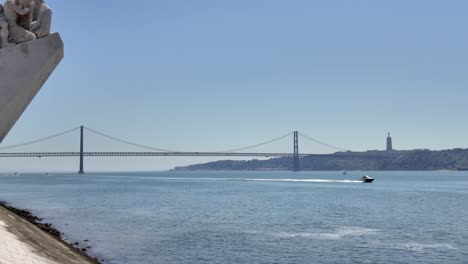 speedboat sailing under the ponte 25 de abril in lisbon, portugal, seen from the monument to the discoveries