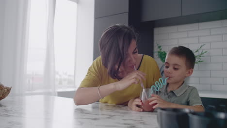 Young-mom-and-son-drinking-juice-with-candy-at-kitchen.-Happy-mother-spending-time-with-son-sitting-on-table-in-slow-motion