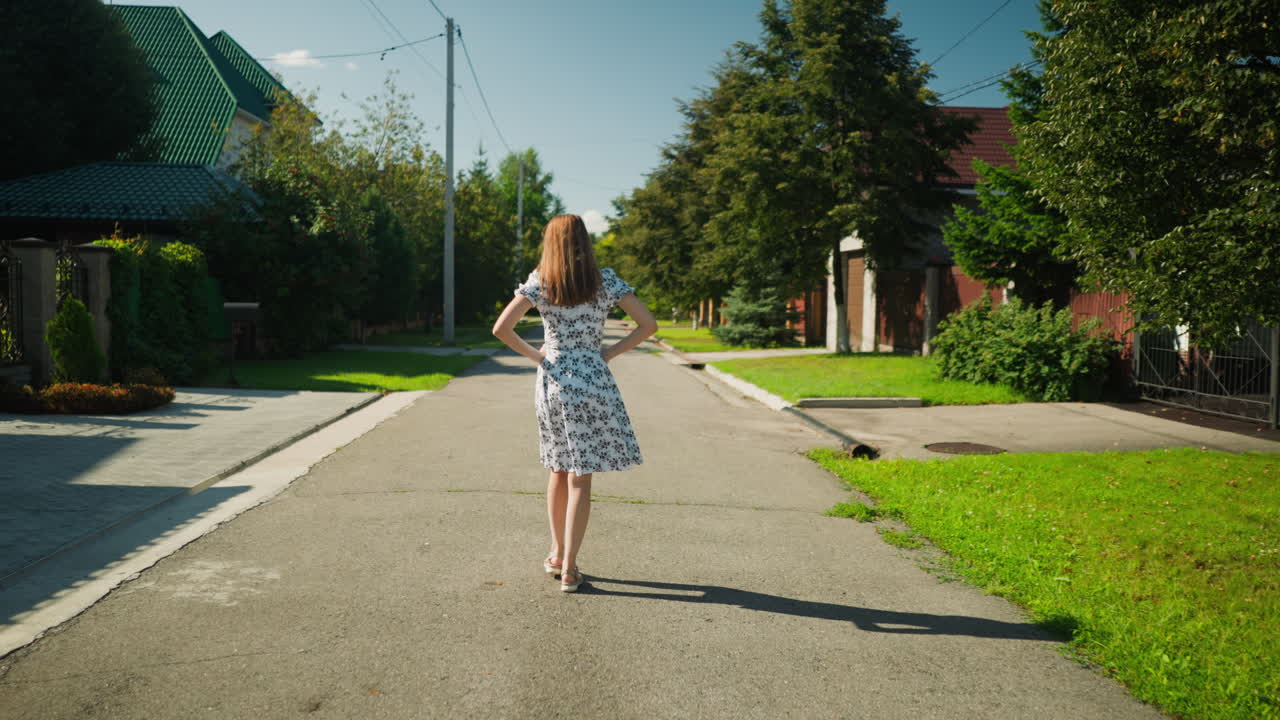Rear view of lady walking with hands on waist along quiet residential street, wearing floral dress, casting strong shadow on sunny day, expressing self assurance and beauty in suburban setting