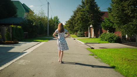 rear view of lady walking with hands on waist along quiet residential street, wearing floral dress, casting strong shadow on sunny day, expressing self assurance and beauty in suburban setting
