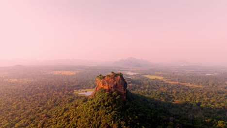 a peaceful sunrise drone shot of sigiriya rock rising above the dense sri lankan jungle, with a calm lake reflecting the first light of day.