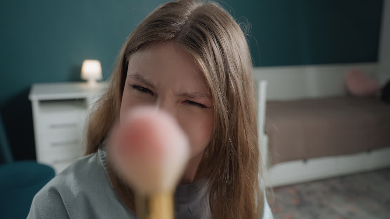 Ragazza adolescente bianca applica il fard con un pennello in una camera da letto accogliente con illuminazione soffusa, pratica una tecnica di trucco naturale, sorride dolcemente, lentiggini visibili, lampada accesa sullo sfondo, primo piano intimo