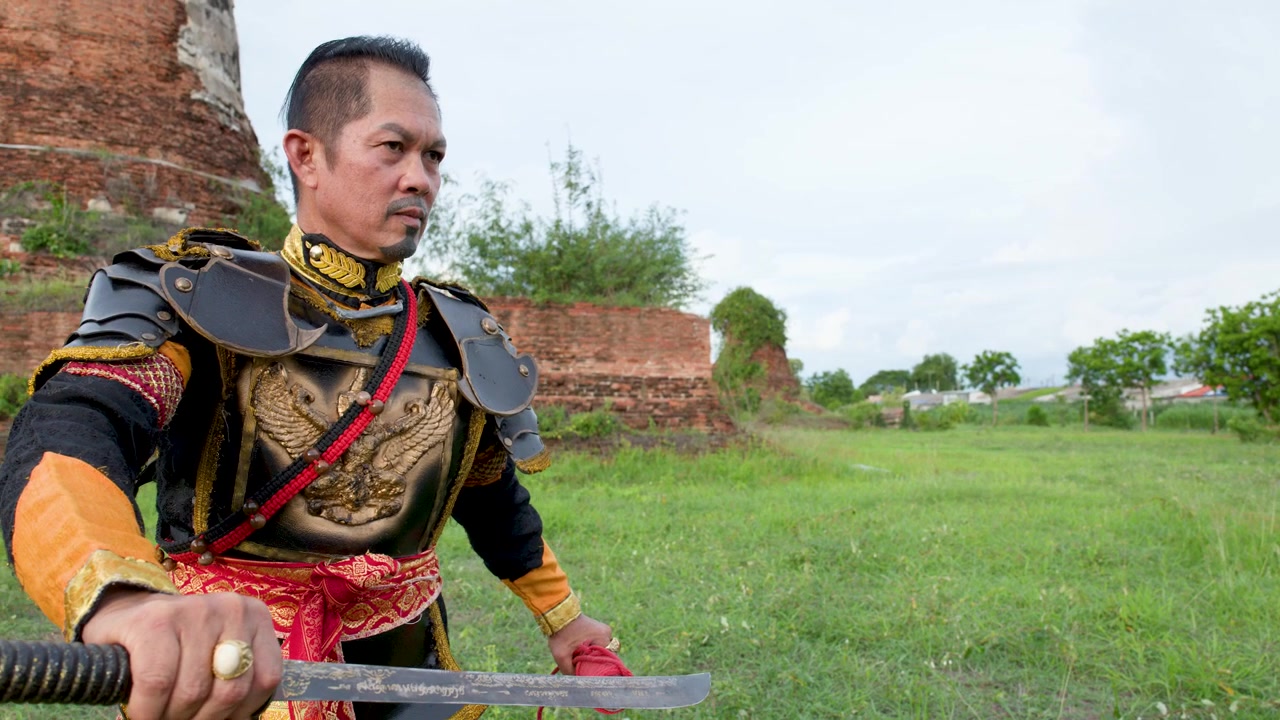 Pria Asia berkostum prajurit bersejarah berdiri di padang rumput dekat reruntuhan bata kuno