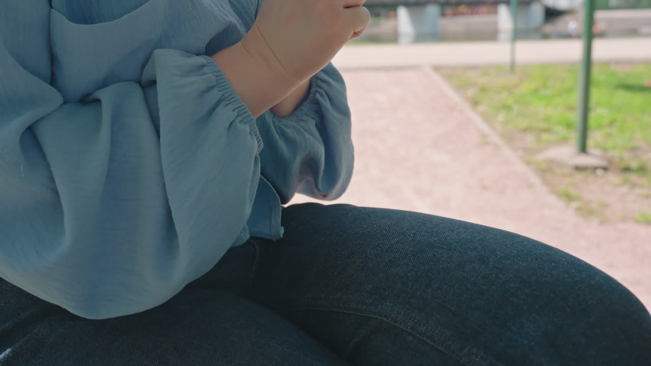 공원에서 청바지를 입고 앉아 두 손으로 무릎을 쓰다듬는 코카서스 인종 여성, 헐렁한 파란색 블라우스 소매, 먼 곳을 응시하며 생각에 잠긴 듯한 모습, 자연광과 벤치 배경이 사색적인 분위기를 강조합니다.