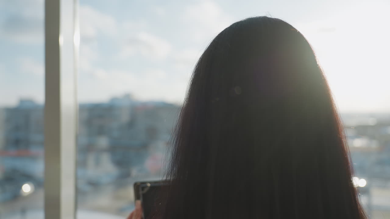 Back view of woman with long straight hair standing indoors using gadget while facing large glass window, bright daylight casting soft glow, urban buildings blurred in background