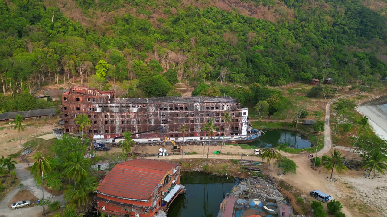 abandoned rusty ghost ship on Koh Chang on tropics island bay, Thailand, during sunset, with a crane working on it. Great aerial view flight static tripod hovering drone