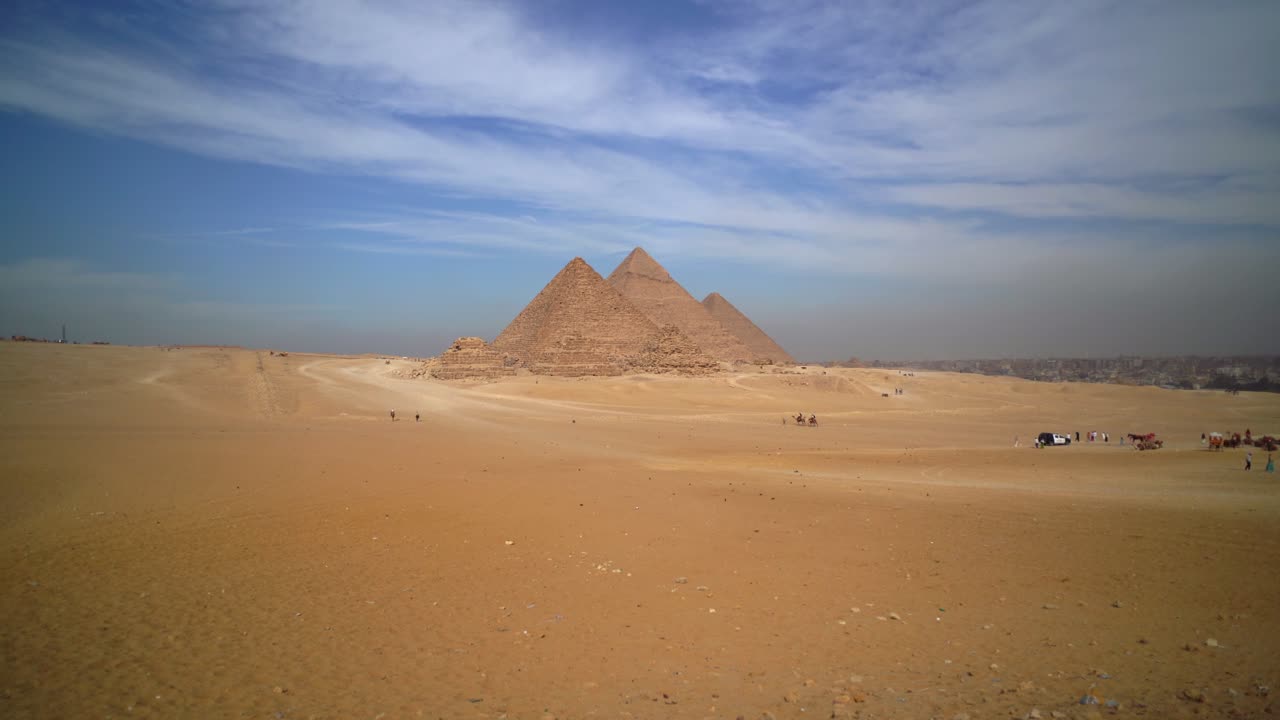 Panorama grandangolare delle tre Piramidi di Giza che sorgono dalle sabbie del deserto in Egitto, con visitatori in lontananza, veicoli turistici e il Cairo visibile sotto nuvole e cielo azzurro.
