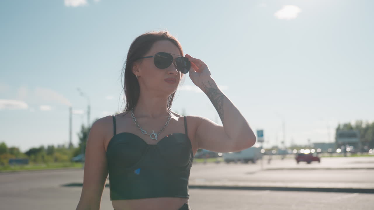 Middle age lady outdoors with tattooed hand and chain necklace in leather crop top holding sunglasses, standing under sunlight with confident expression and blurred urban background