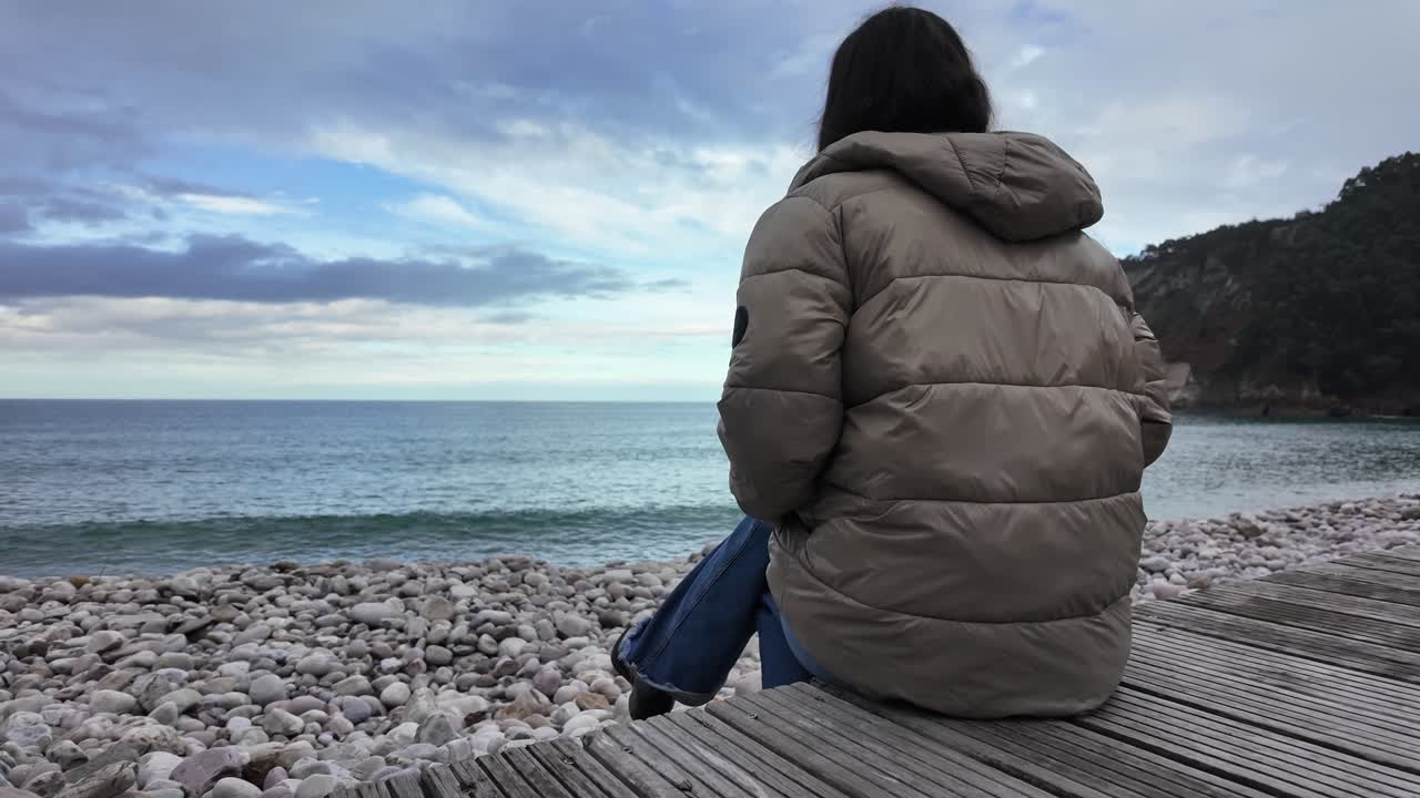 Slow-motion view of a woman seated on seaside rocks under a cloudy sky