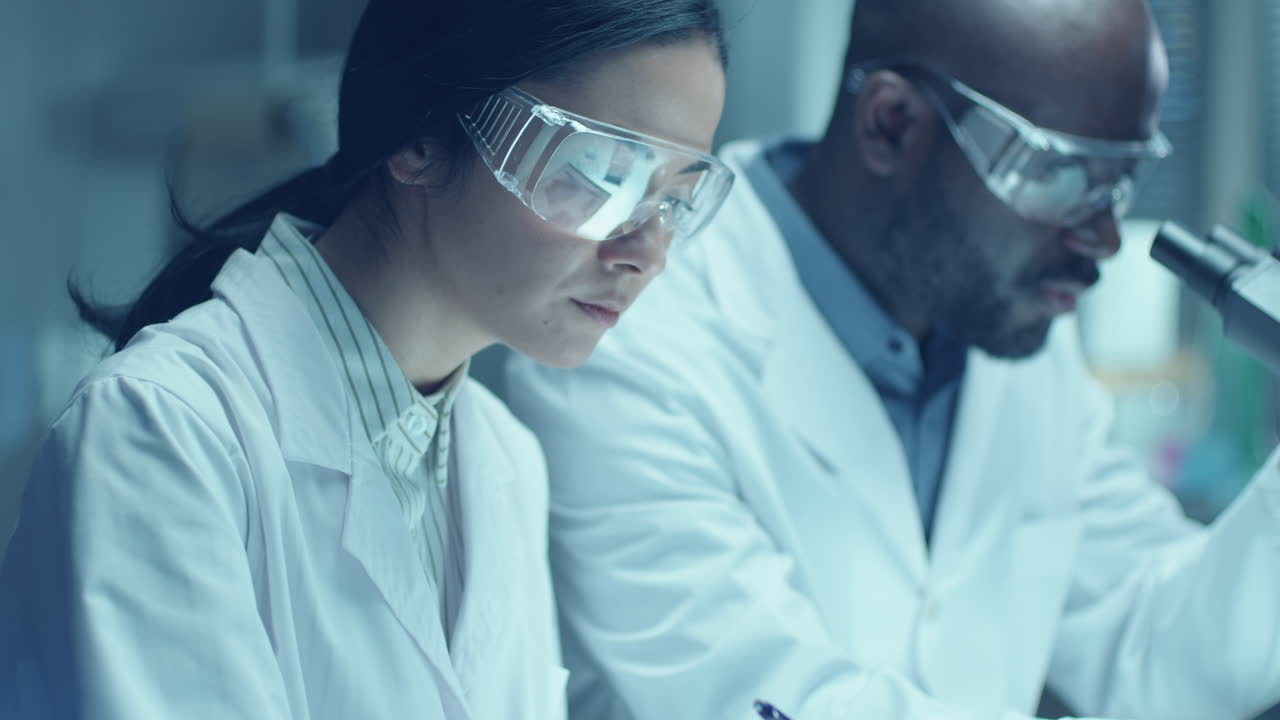 Black Chemist Doing Laboratory Research with Female Assistant Taking Notes