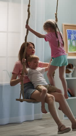 family relaxes on swing. woman with kids play in room with sofa and toys. toddler boy sits on mother lap and preschooler girl stands looking at brother contentedly