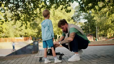 Dad-helps-his-little-son-put-on-protective-clothing-before-riding-a-skateboard-at-the-skatepark-in-the-park.-Father-and-son-lead-an-active-lifestyle