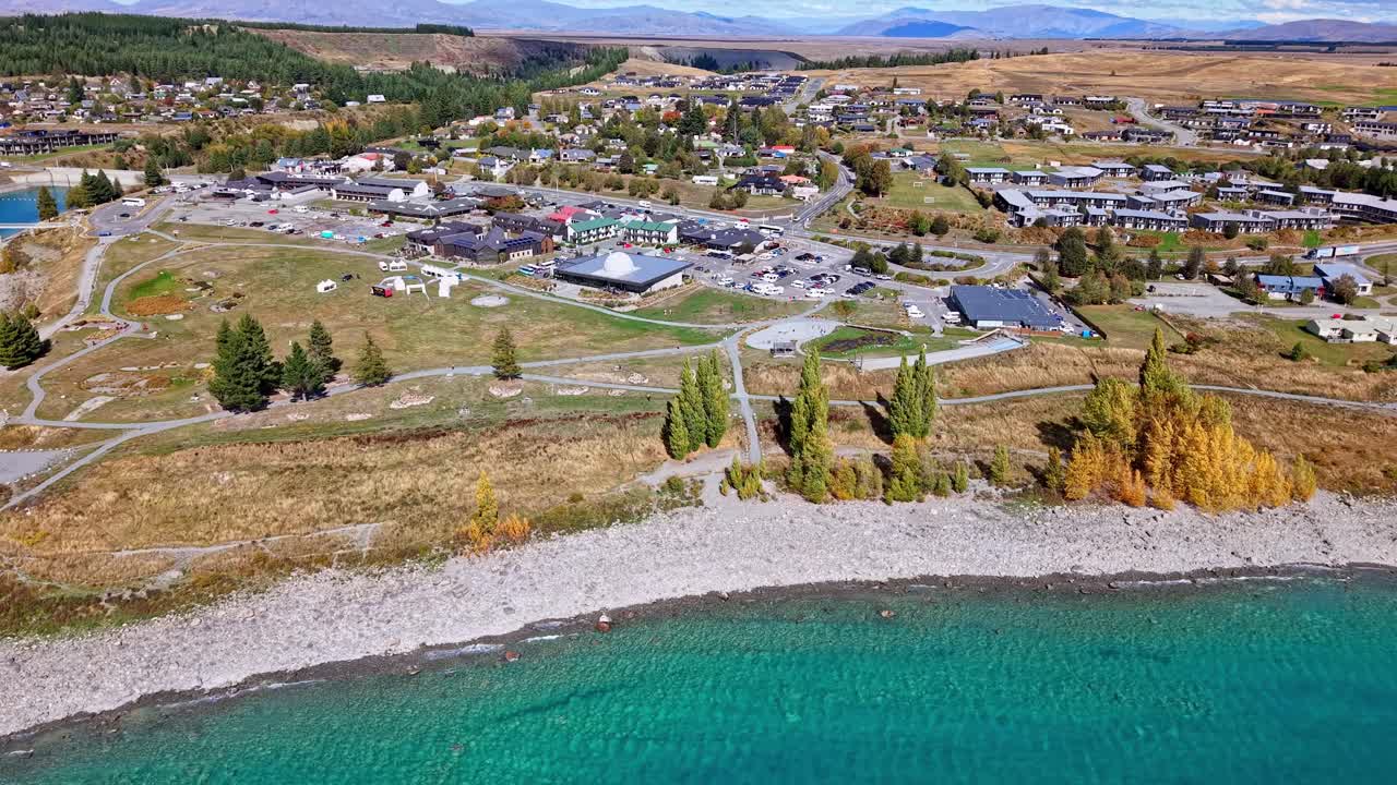 Drone pulls back over Tekapo town showing Mt John Observatory, roads, and houses before revealing the turquoise lake and distant mountains