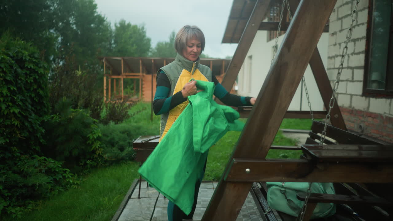 mujer de mediana edad en chaqueta amarilla y verde con botas amarillas se encuentra cerca del banco de columpio, desplegando un impermeable verde, el fondo presenta vegetación exuberante, árboles y jardín colorido