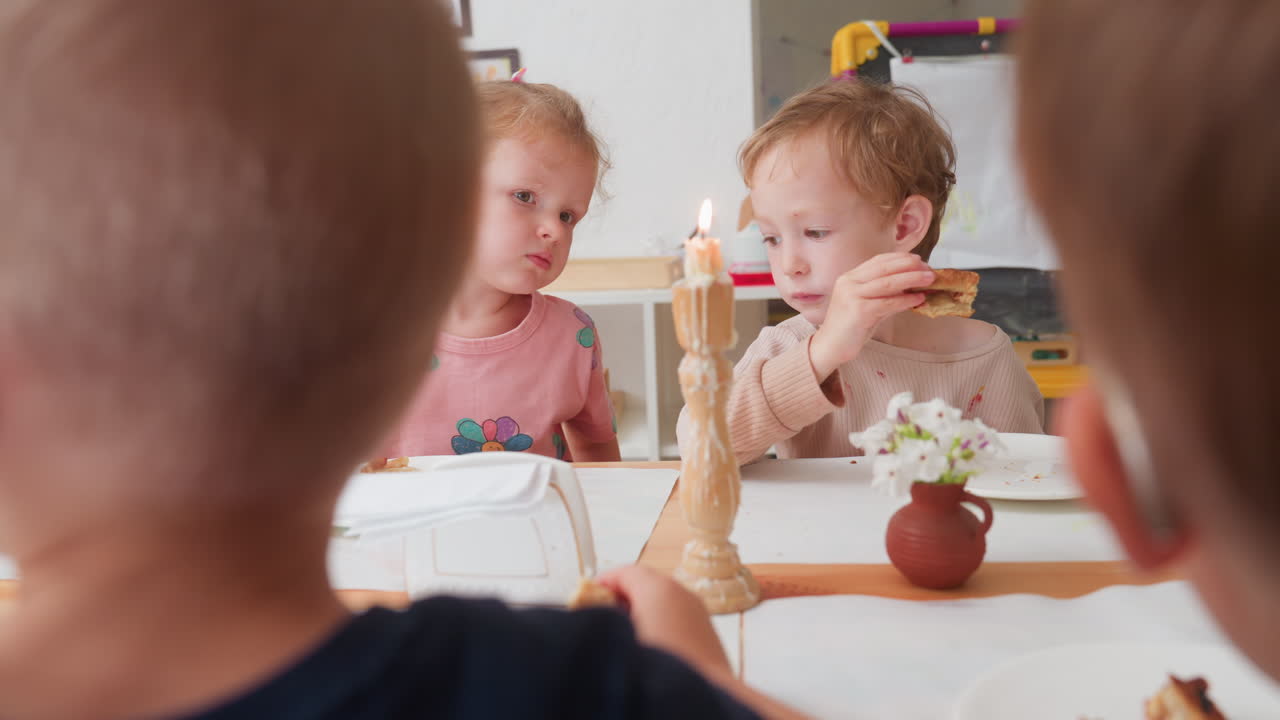 간식 테이블에 앉아 있는 유치원 아이들, 핑크 꽃무늬 셔츠를 입은 여자아이가 이야기를 나누고 남자아이는 빵을 들고 있어요. 촛불이 켜져 있고, 반 친구들은 흐릿하게 보이며, 아늑한 교실 분위기, 함께하는 식사, 그리고 호기심이 느껴져요.