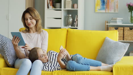 Caucasian-young-mother-and-teen-daughter-resting-on-the-sofa-at-home-and-using-the-tablet-device-and-smartphone.-Indoors.