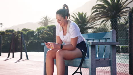 woman sitting on a bench on a tennis court using her phone