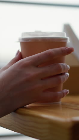 primer plano de la mano de la dama tocando la taza de café en la mesa de madera con fondo borroso suave, vista parcial del teléfono visible, entorno de café moderno con luz natural, momento de relajación con bebida caliente