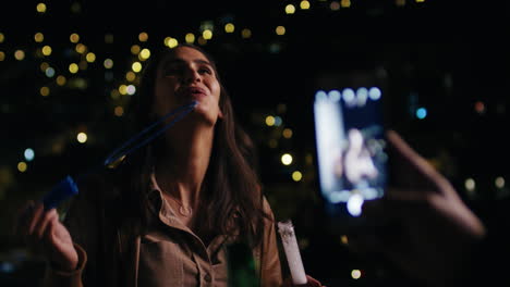 Hermosa-Joven-Soplando-Burbujas-A-Su-Novio-Usando-Un-Teléfono-Inteligente-Tomando-Fotos-En-La-Azotea-Por-La-Noche-Compartiendo-Una-Velada-Romántica-Con-Luces-Bokeh-De-La-Ciudad-En-El-Fondo