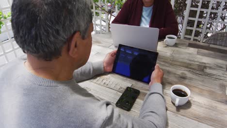 Senior-mixed-race-couple-sitting-in-garden-using-laptop-and-tablet