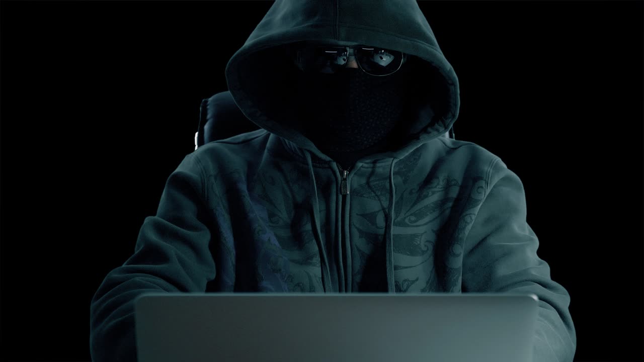 hombre con capucha sentado en la mesa y codificando en el portátil. vista frontal de cerca. hacker con máscara de balaclava y gafas trabajando en la computadora. navegando por internet hombre con fondo de hormigón. clave de croma del canal alfa.