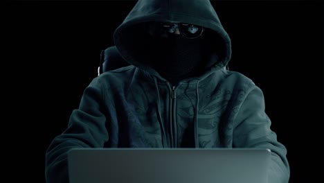 man in hood sitting at table and coding on laptop. front view close up. hacker in balaclava mask and glasses working at computer. surfing internet man at concrete background. alpha channel chroma key.