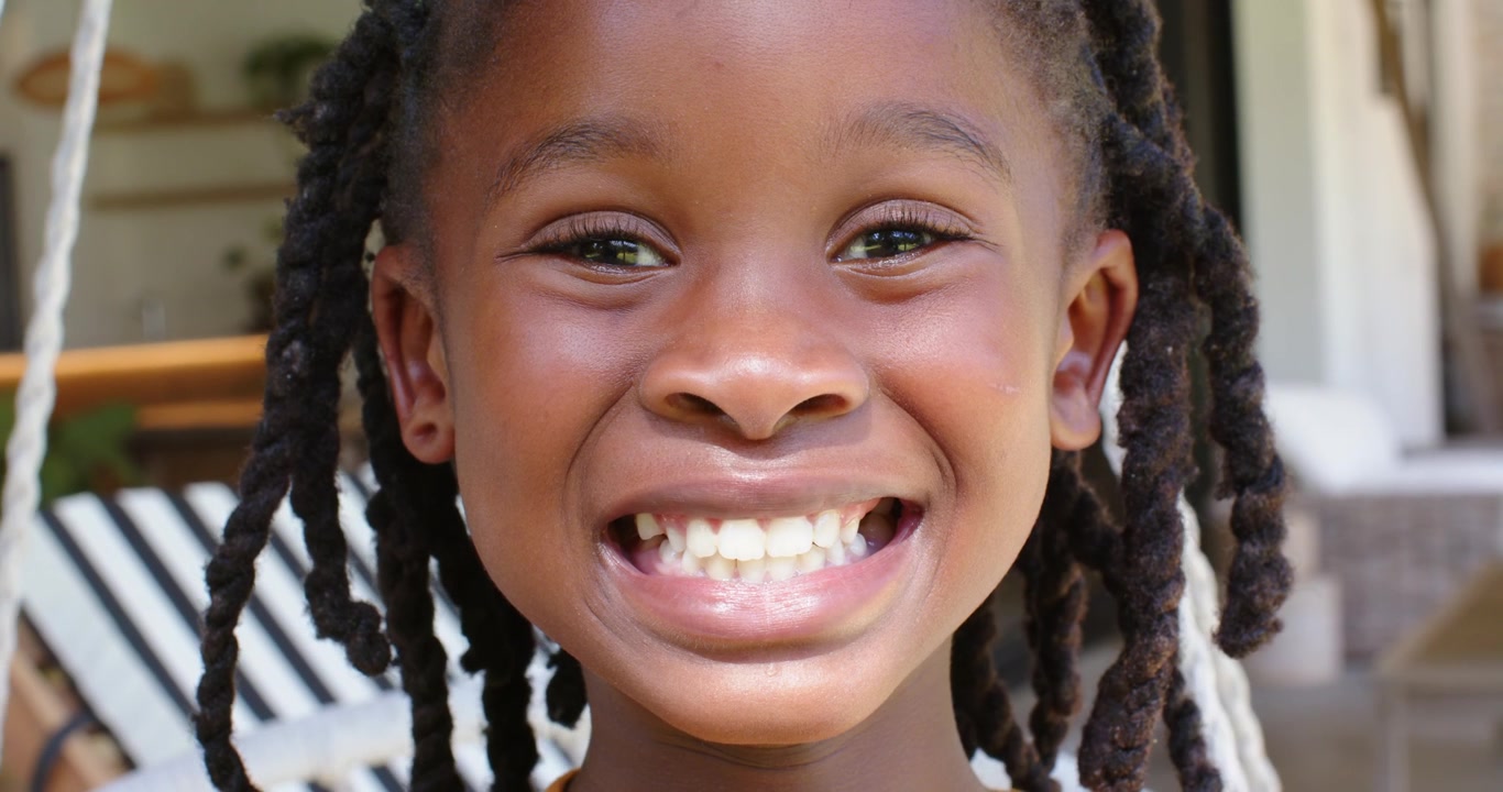 Bambino africano che sorride mentre lo sguardo della telecamera allarga il sorriso sull'altalena del portico, con le trecce che ondeggiano e un top color senape.
