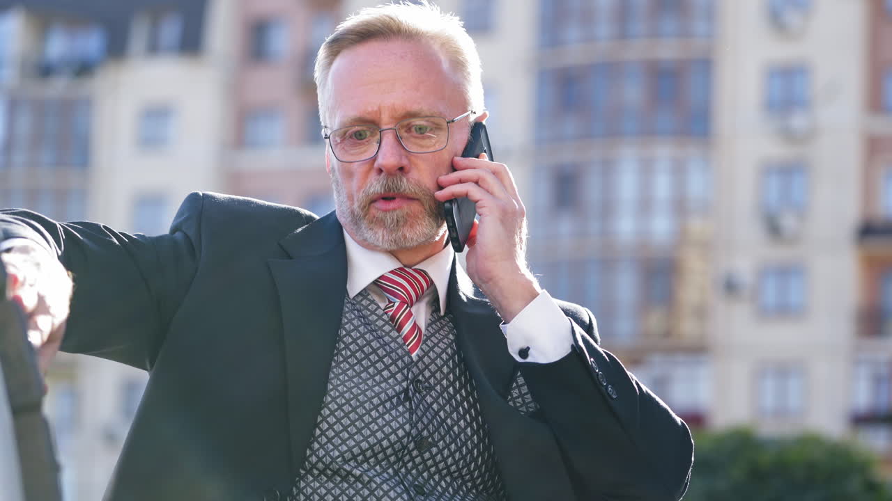 Portrait of successful senior businessman using cell phone while sitting on a bench. Mature man answering phone call and explaining business technology on mobile phone in city.