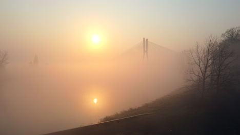 Low-flying-over-a-wide-river,-towards-a-modern-cable-bridge-during-a-foggy-sunrise-morning