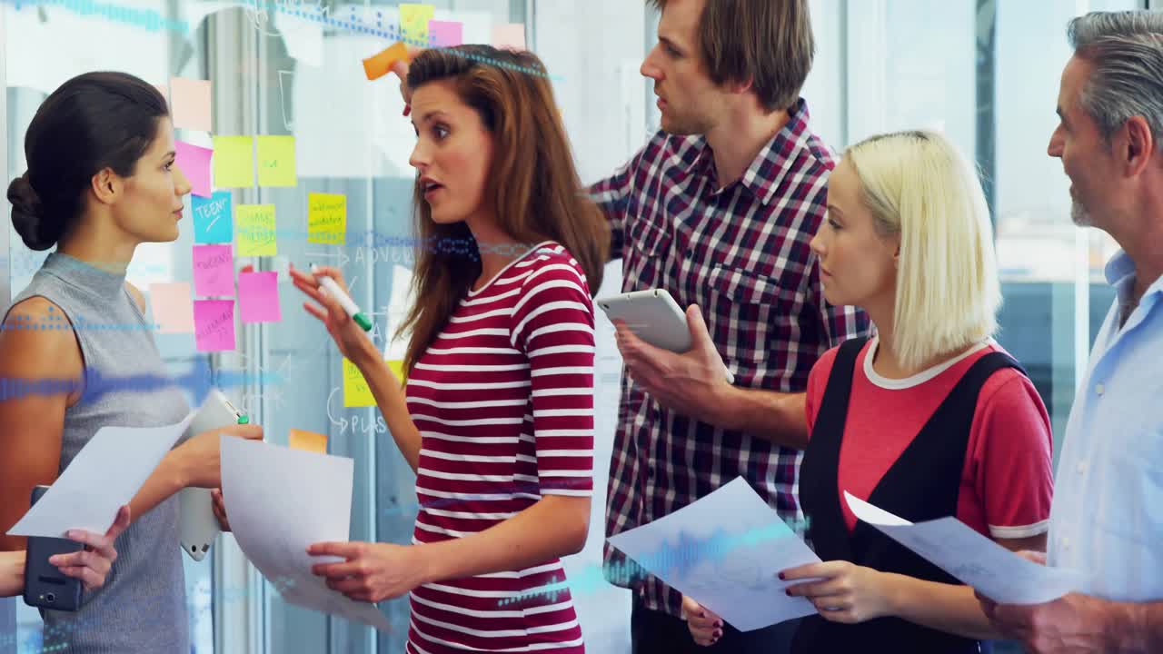 Colleagues brainstorming around glass partition using sticky notes, tablets in business office