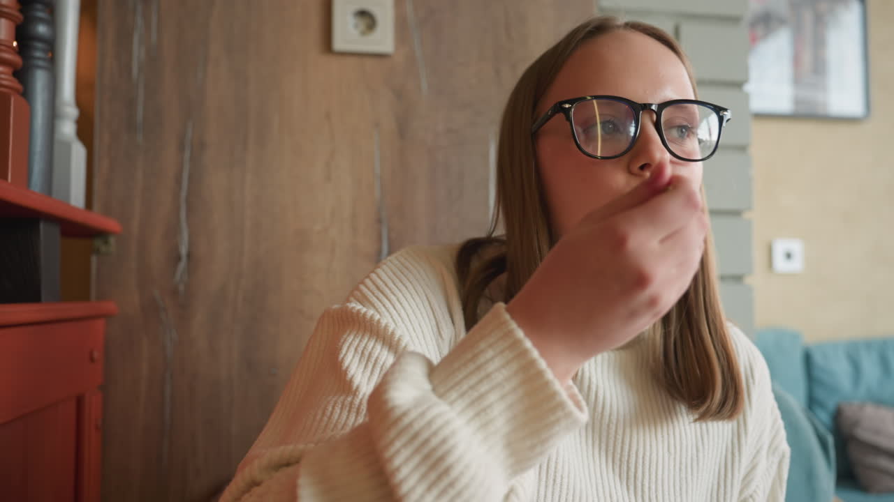 Young girl in glasses and white sweater sits in cozy cafe enjoying piece of cake while seated near wooden wall and blue sofa with blurred background