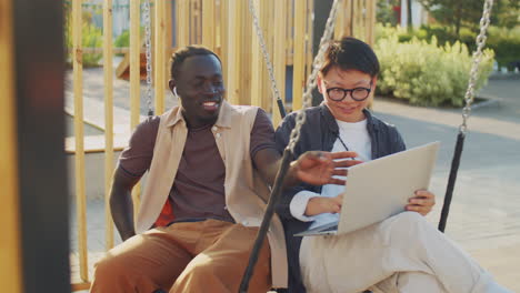 black man and asian woman using laptop and speaking on swing