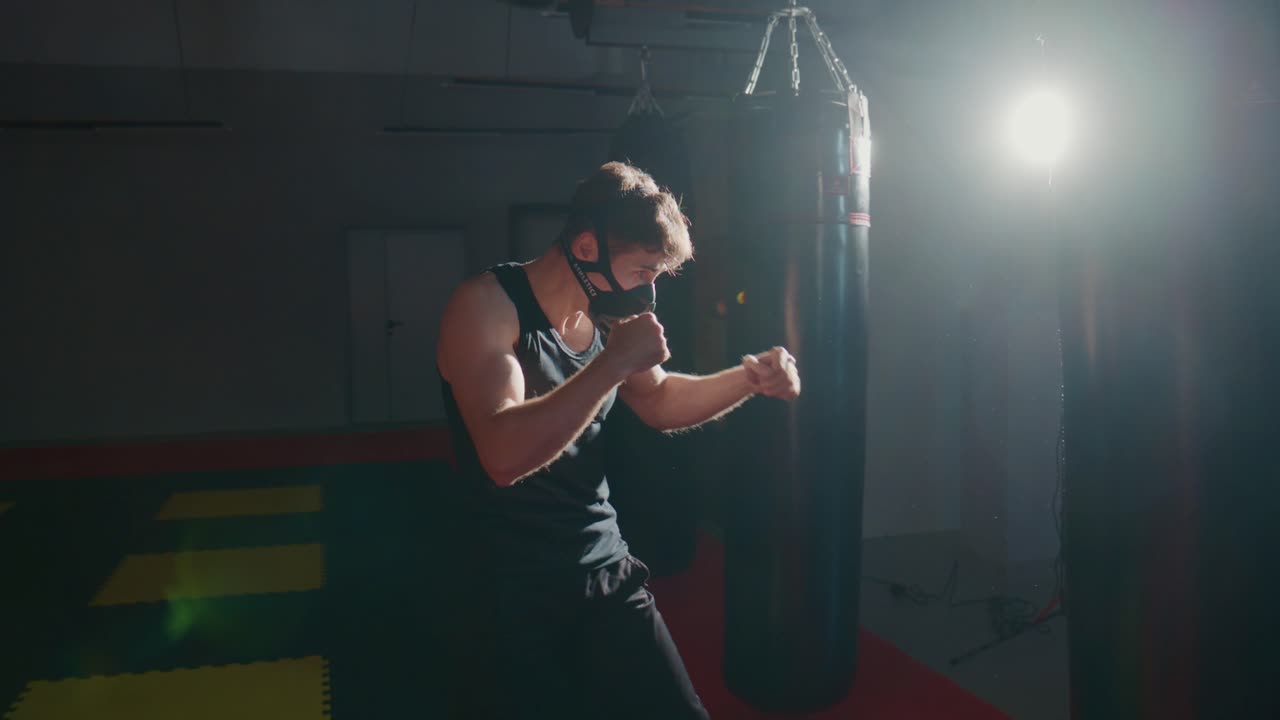 Young Man in Training Mask Boxing Punching Bag in Dark Gym
