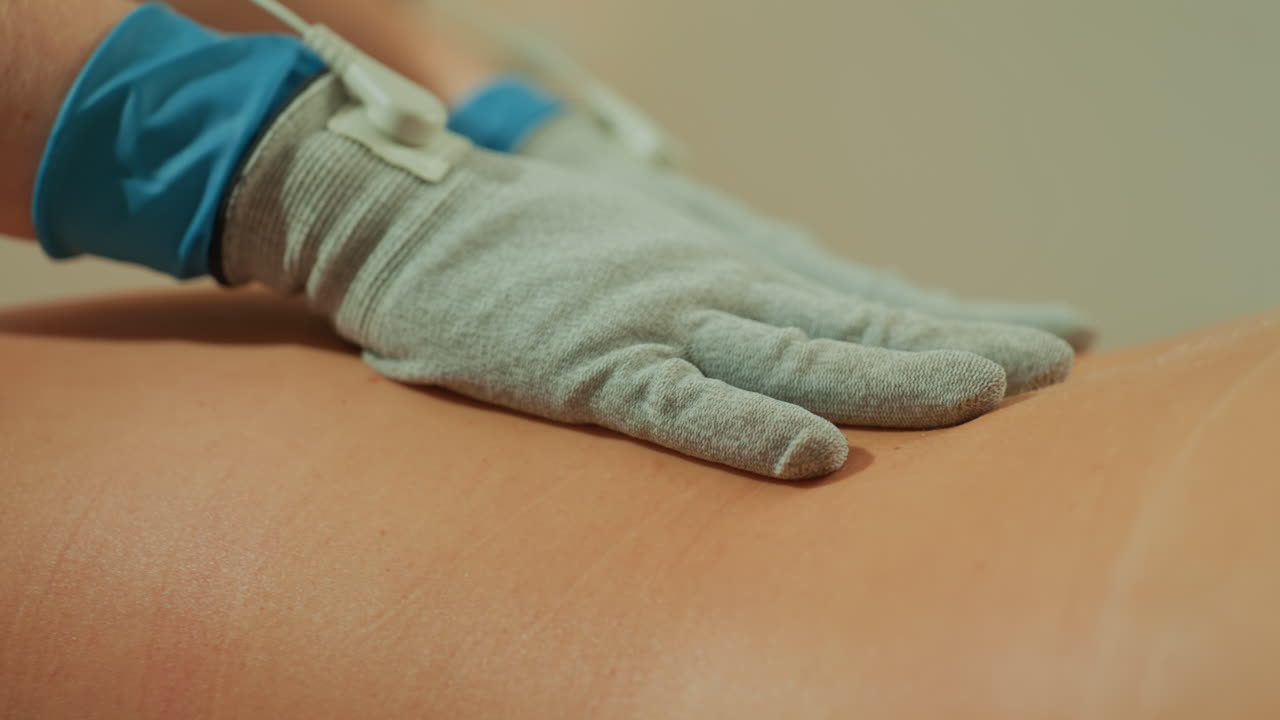 Donna caucasica sottoposta a terapia a microcorrente con guanti, clinico che applica un massaggio con elettrodi pulsati con posizionamento preciso delle mani su lombare e collo, primo piano del movimento del guanto conduttivo, concentrata.