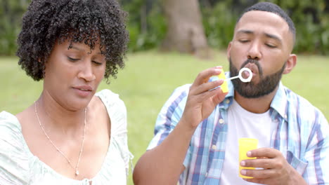 happy african american couple blowing bubbles sitting in garden, slow motion