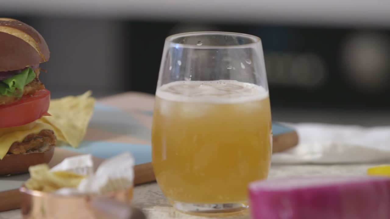A vibrant homemade chicken burger stacked with fresh vegetables and cheese is paired with a cold drink and tortilla chips, captured in a close-up with natural light.