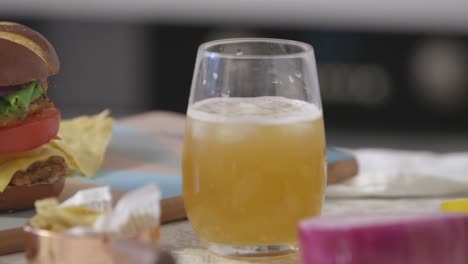 a vibrant homemade chicken burger stacked with fresh vegetables and cheese is paired with a cold drink and tortilla chips, captured in a close-up with natural light.