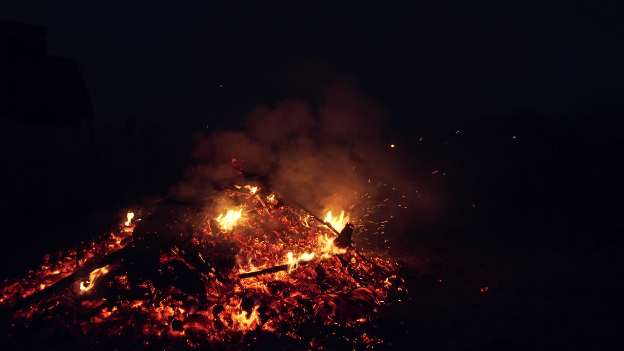 Burning red hot sparks rise from large fire in the night sky. Smoldering ashes from a large fire. A man turns over logs on fire with a pitchfork