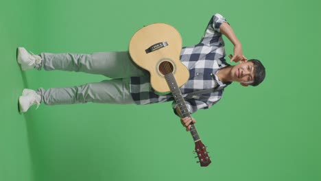 full body of asian teenager with guitar showing call me gesture and smiling on green screen background