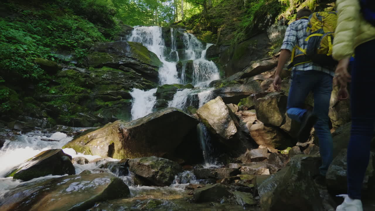 un par de viajeros con mochilas escalando rocas hasta una hermosa cascada de video de 4k y 10 bits