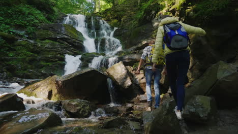 un par de viajeros con mochilas escalando rocas hasta una hermosa cascada de video de 4k y 10 bits