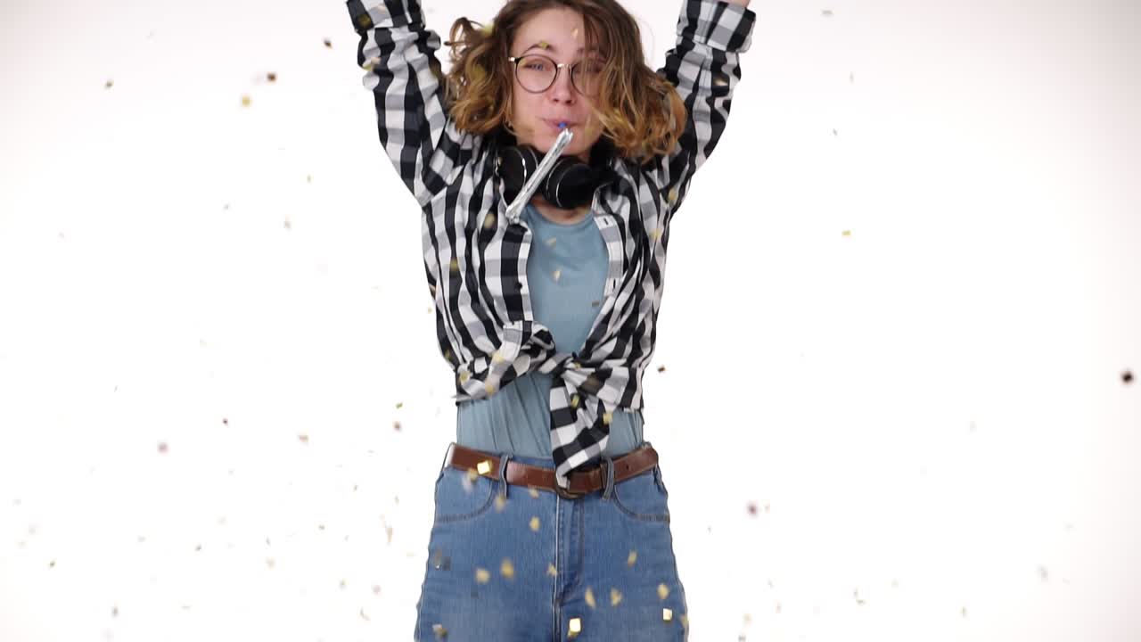 Portrait of excited young woman in plaid shirt and headphones on neck in birthday colorful hat blow in pipe, falling confetti isolated over white background in studio. People sincere emotions, holiday lifestyle concept. Celebrating party day