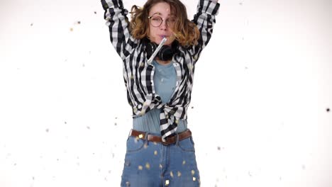 Portrait-of-excited-young-woman-in-plaid-shirt-and-headphones-on-neck-in-birthday-colorful-hat-blow-in-pipe,-falling-confetti-isolated-over-white-background-in-studio.-People-sincere-emotions,-holiday-lifestyle-concept.-Celebrating-party-day