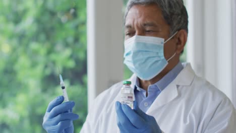 African-american-male-senior-doctor-wearing-face-mask-holding-a-syringe-and-covid-19-vaccine