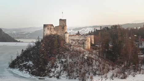 Idílico-Castillo-Niedzica-Al-Atardecer-En-La-Cima-De-Una-Colina-En-El-Lago-Congelado-Czorsztyn,-Antena