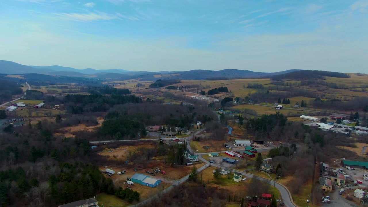 Droneskud, der stiger op over en landlig bjergby omgivet af bølgende bakker og skov i dagslys i Stanford, New York.