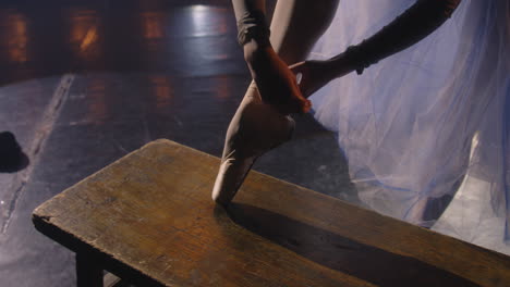 beautiful ballet female dancer in dress puts on and ties pointe shoes for choreography rehearsal. ballerina practices on theater stage illuminated by spotlights. classical ballet dance performance.