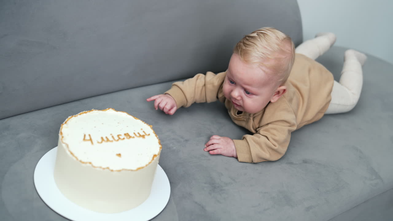 Crying little blond baby lies on the sofa. Big white cake with 4 months sign is in front of a kid.
