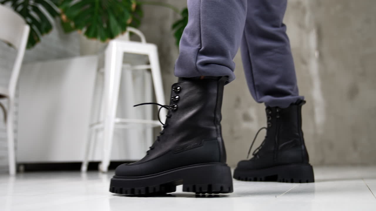 Black laced army boots demonstration. Close up. Model wearing grey pants and leather massive footwear in studio.