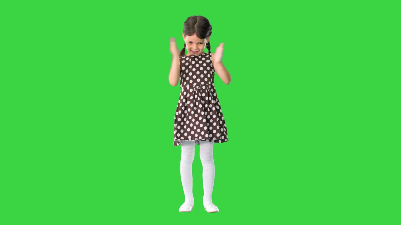una niña sonriente con un vestido de puntos polca aplaudiendo sus manos vigorosamente mirando a la cámara en una pantalla verde, clave de croma